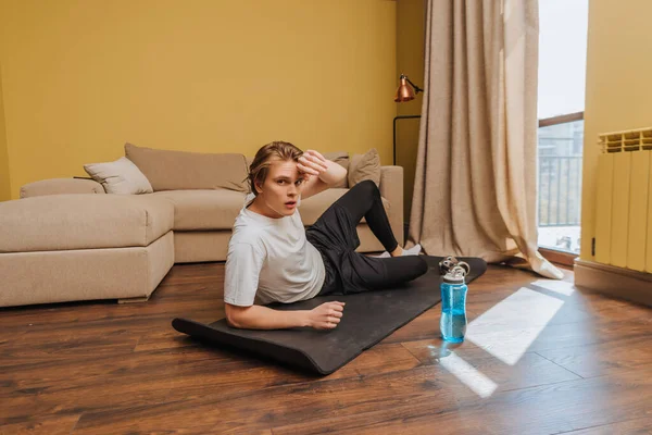 Homem cansado deitado no tapete de fitness perto de garrafa de esportes com água, fim do conceito de quarentena — Fotografia de Stock