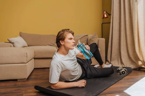 Homem bonito deitado no tapete de fitness e segurando garrafa de esportes com água, fim do conceito de quarentena — Fotografia de Stock