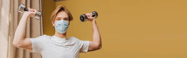 Orientación panorámica del hombre en máscara médica ejercicio con pesas en la sala de estar - foto de stock