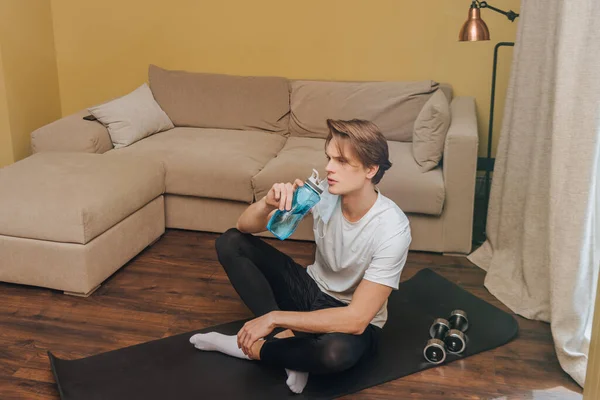 Bel homme assis sur un tapis de fitness et tenant une bouteille de sport avec de l'eau, concept de fin de quarantaine — Photo de stock