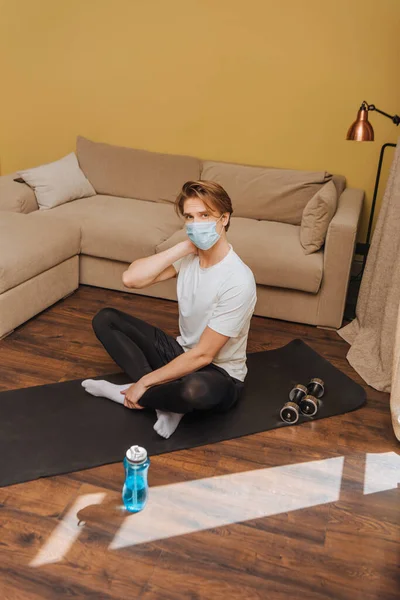 Uomo sportivo in maschera medica seduto sul tappetino fitness vicino bottiglia sportiva con acqua — Foto stock