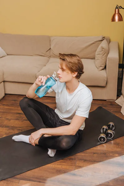 Hombre guapo sentado en la alfombra de fitness y agua potable, fin del concepto de cuarentena - foto de stock