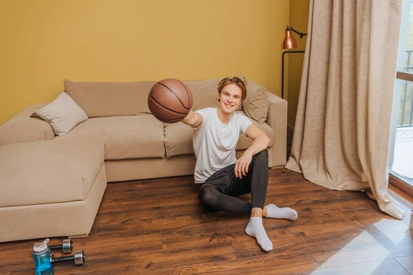 Happy man holding football while sitting on floor near sports bottle and dumbbells, end of quarantine concept — Stock Photo