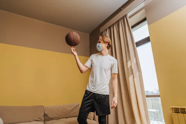 Man in medical mask spinning basketball on finger at home — Stock Photo