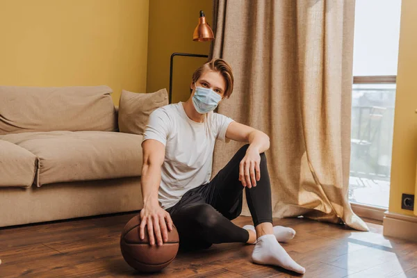 Hombre con máscara médica sentado en el suelo con baloncesto - foto de stock