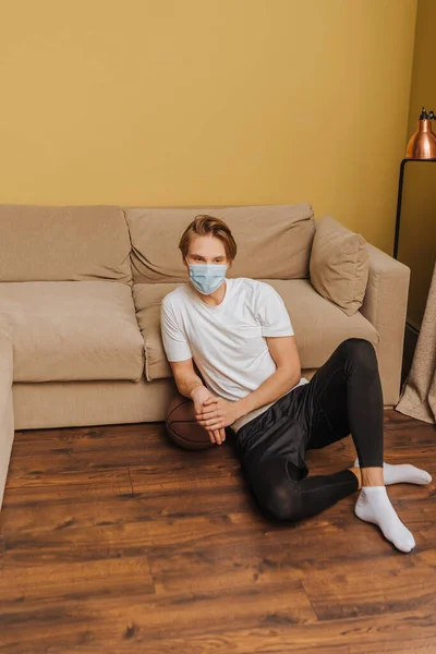 Mann mit medizinischer Maske sitzt auf dem Boden neben Basketball und Sofa — Stockfoto