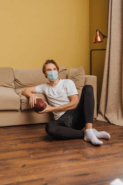 Joven en máscara médica sosteniendo fútbol americano y sentado en el suelo en la sala de estar - foto de stock