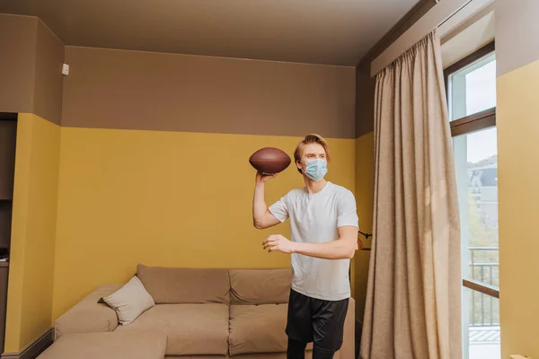 Mann mit medizinischer Maske hält American Football im Wohnzimmer — Stockfoto