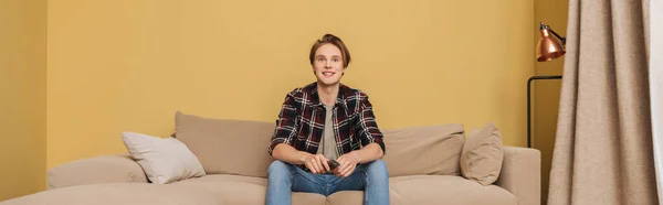 Panoramic crop of happy man holding remote controller and sitting on sofa, end of quarantine concept — Stock Photo