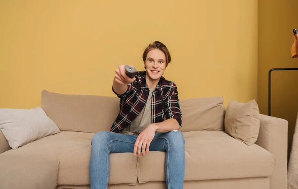 Joven sonriente hombre sosteniendo mando a distancia y sentado en el sofá, fin del concepto de cuarentena - foto de stock