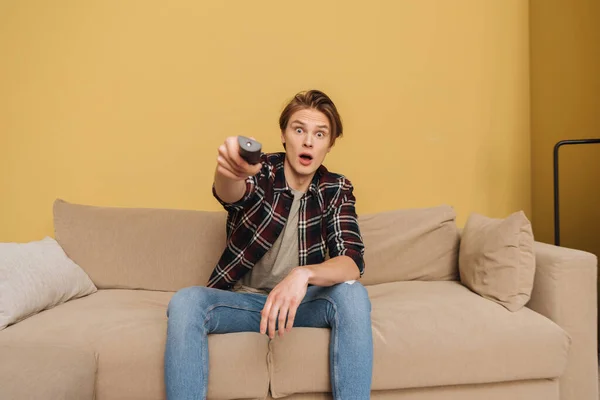 Selective focus of surprised man holding remote controller and sitting on sofa, end of quarantine concept — Stock Photo