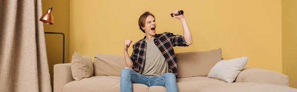 Panoramic orientation of excited man with opened mouth sitting on sofa and holding remote controller, end of quarantine concept — Stock Photo