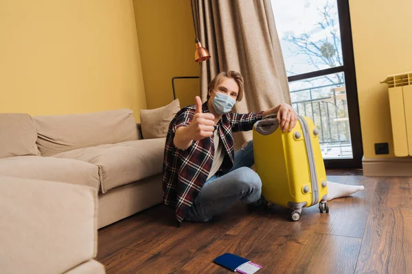 Hombre con máscara médica mostrando el pulgar hacia arriba cerca del pasaporte con billete de avión y equipaje, fin del concepto de cuarentena - foto de stock