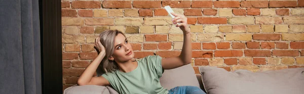 Foto panorámica de la hermosa mujer joven tomando selfie en el teléfono inteligente mientras se relaja en el sofá - foto de stock