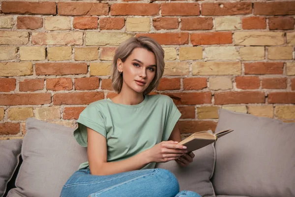 Attraktive junge Frau blickt in die Kamera, während sie mit aufgeschlagenem Buch auf dem Sofa sitzt — Stockfoto