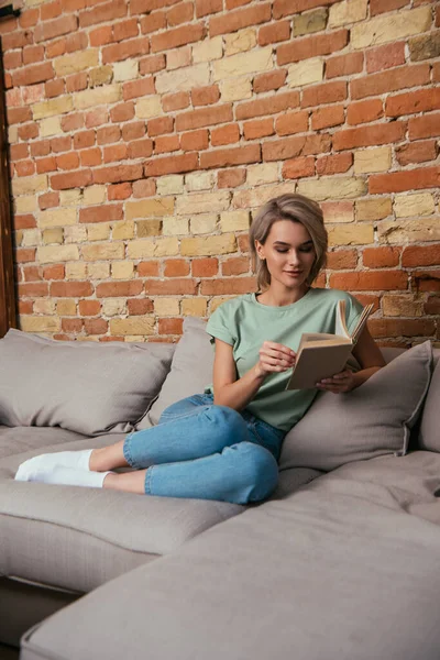 Bella giovane donna lettura libro mentre seduto sul divano vicino muro di mattoni — Foto stock