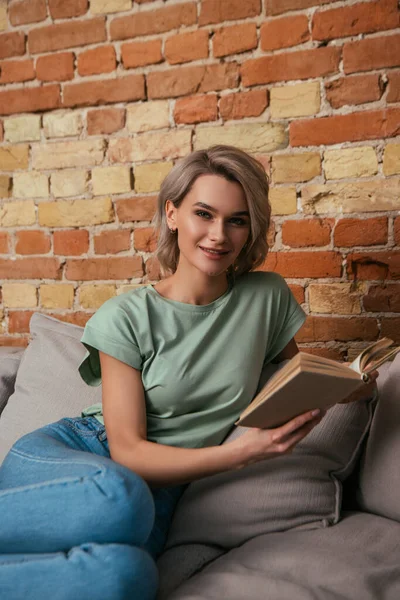Attractive young woman smiling at camera while sitting on sofa with open book — Stock Photo