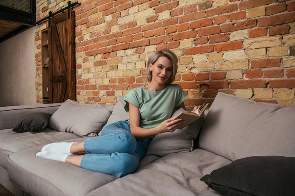 Lächelnde junge Frau blickt in die Kamera, während sie es sich auf dem Sofa mit Buch gemütlich macht — Stockfoto
