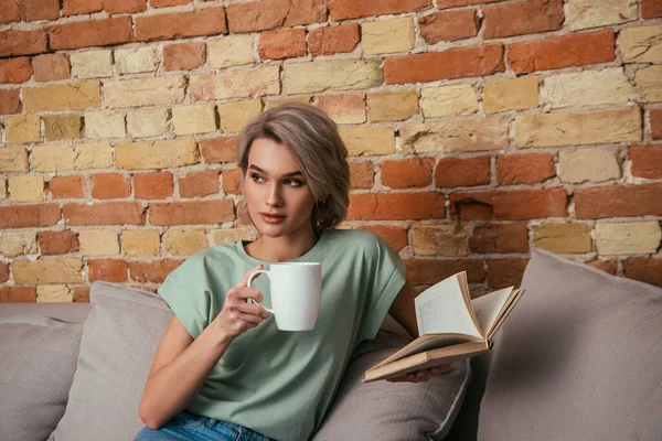 Chère jeune femme tenant tasse de thé et livre ouvert tout en étant assis sur le canapé près du mur de briques — Photo de stock