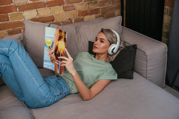 High angle view of young woman in wireless headphones reading beauty and style journal while relaxing on sofa at home — Stock Photo