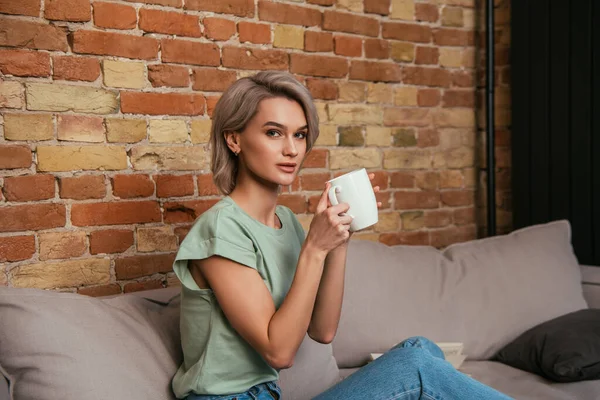 Beau jeune homme regardant la caméra tout en étant assis sur le canapé et tenant une tasse de thé — Photo de stock