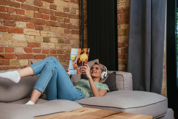Donna sorridente in cuffie wireless che legge bellezza e stile rivista mentre sdraiato sul divano a casa — Foto stock