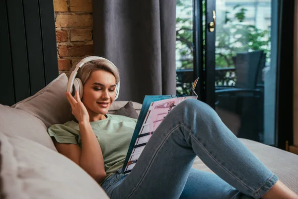 Smiling woman in wireless headphones reading magazine while resting on sofa at home — Stock Photo