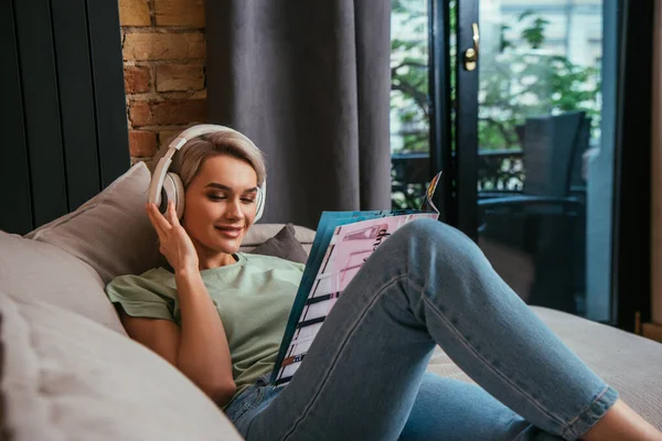 Femme souriante dans un casque sans fil lecture magazine tout en se reposant sur le canapé à la maison — Photo de stock