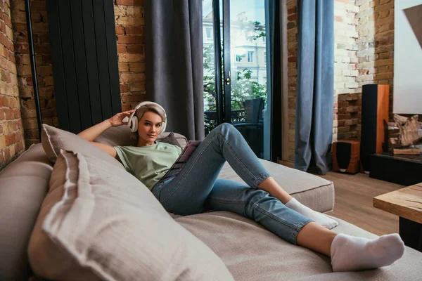Selektive Fokussierung der schönen Frau in drahtlosen Kopfhörern, die auf dem Sofa in die Kamera schaut — Stockfoto