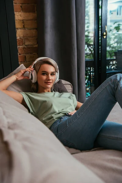 Enfoque selectivo de mujer atractiva en auriculares inalámbricos sonriendo a la cámara mientras se relaja en el sofá - foto de stock