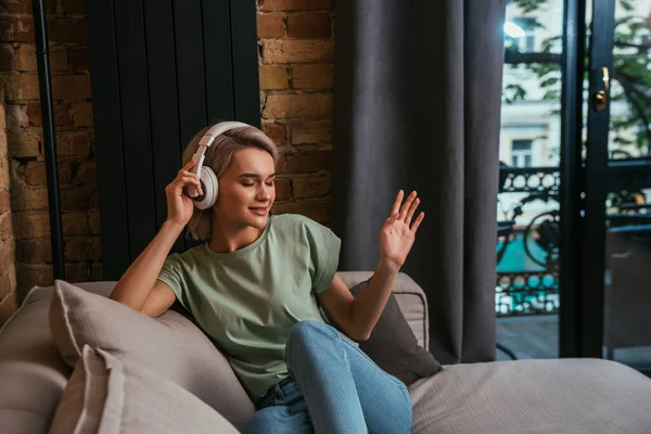 Mulher sorrindo gesticulando enquanto sentada no sofá e ouvindo música em fones de ouvido sem fio com olhos fechados — Fotografia de Stock