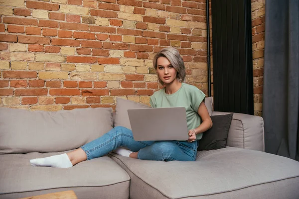 Schöne junge Frau blickt in die Kamera, während sie mit Laptop auf dem Sofa sitzt — Stock Photo