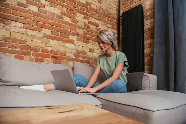Attractive young woman using laptop while resting on sofa near brick wall at home — Stock Photo
