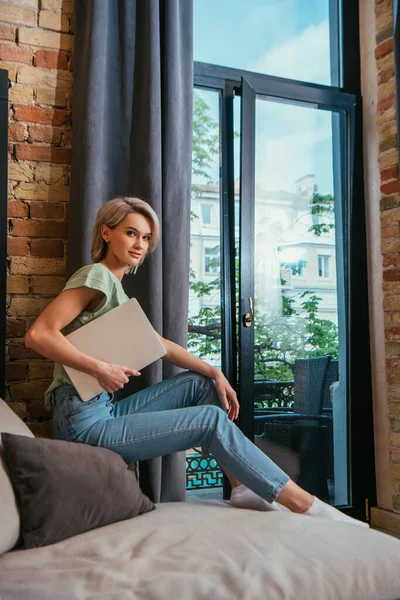 Lächelnd blickt eine junge Frau in die Kamera, während sie auf einem Sofa am Fenster sitzt und Laptop in der Hand hält — Stockfoto