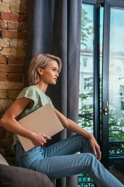 Vista lateral de la mujer pensativa sosteniendo el ordenador portátil mientras está sentado cerca de la ventana en casa - foto de stock