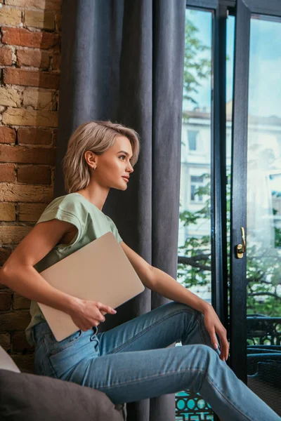 Vista laterale della donna pensierosa che tiene il computer portatile mentre si siede vicino alla finestra a casa — Foto stock