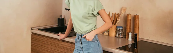 Vista recortada de la mujer sosteniendo la mano en el bolsillo mientras está de pie en la cocina, tiro panorámico - foto de stock