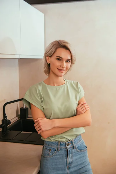 Lächelnde, schöne Frau, die mit verschränkten Armen in der Küche steht und in die Kamera schaut — Stockfoto