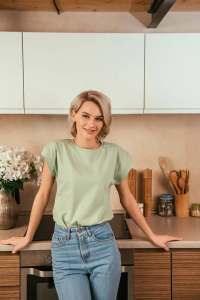 Glückliche, attraktive Frau lächelt in die Kamera, während sie in der Küche steht — Stockfoto