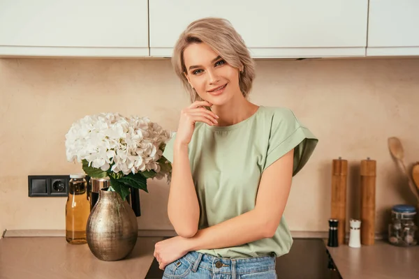 Felice, bella donna che tocca il viso mentre guarda la fotocamera in cucina — Foto stock