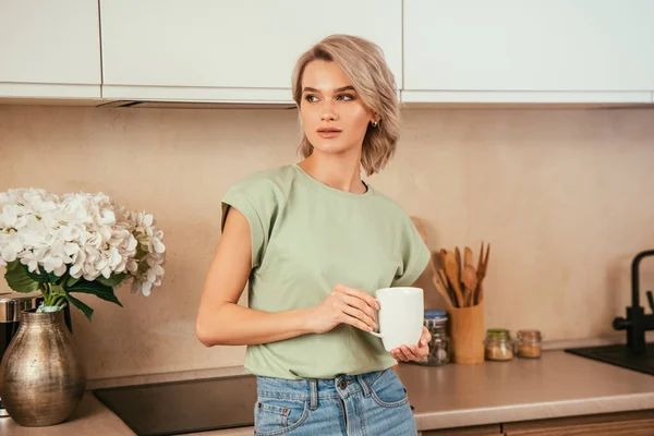 Giovane donna premurosa che guarda lontano mentre tiene in mano una tazza di tè in cucina — Foto stock