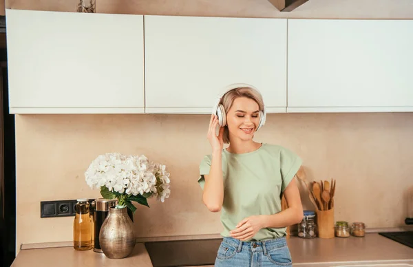 Feliz joven escuchando música en auriculares inalámbricos en la cocina con los ojos cerrados - foto de stock