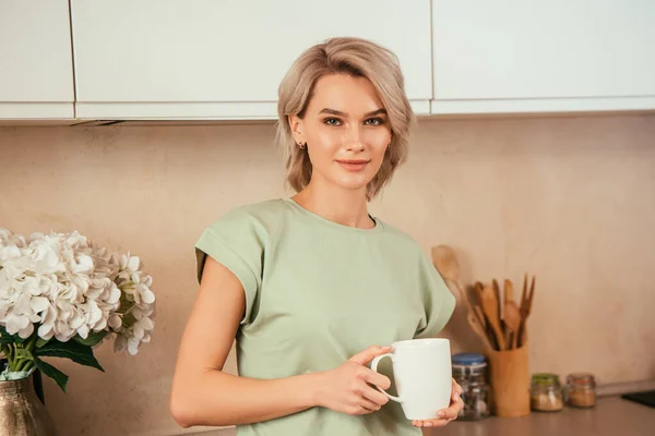 Selbstbewusste, attraktive Frau, die in die Kamera schaut, während sie in der Küche Tee trinkt — Stockfoto