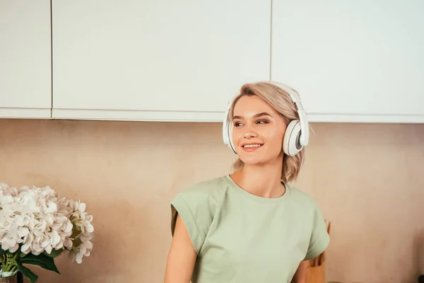 Souriant jeune femme écouter de la musique dans les écouteurs et regarder loin dans la cuisine — Photo de stock