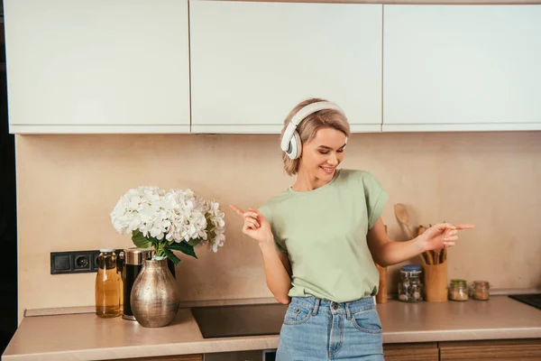 Happy young woman gesturing while listening music in wireless headphones in kitchen — Stock Photo