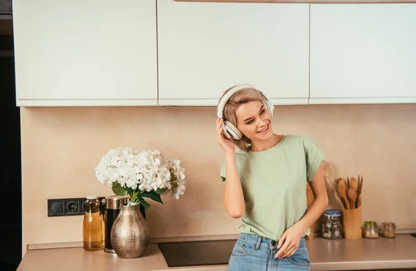 Sorrindo, mulher atraente ouvir música em fones de ouvido sem fio na cozinha — Fotografia de Stock