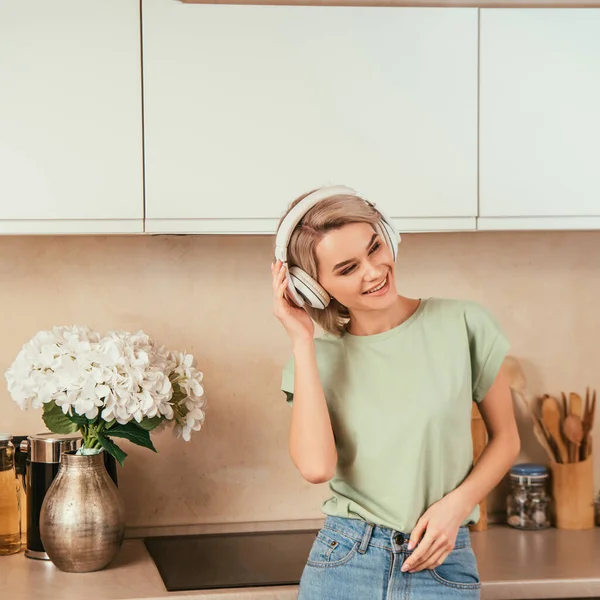 Happy young woman listening music in wireless headphones in kitchen — Stock Photo