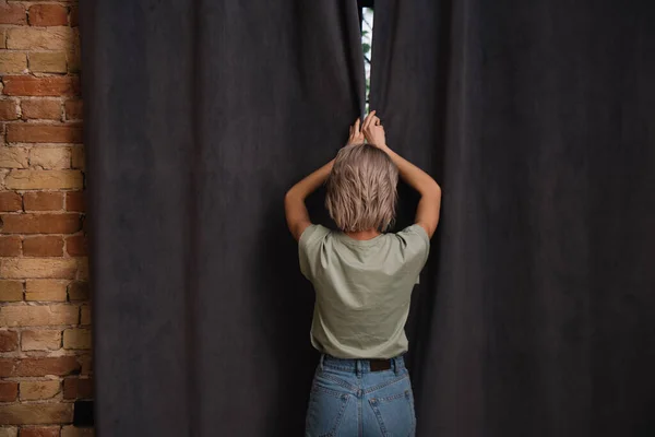 Vue arrière de la jeune femme ouvrant des rideaux sombres sur la fenêtre à la maison — Photo de stock