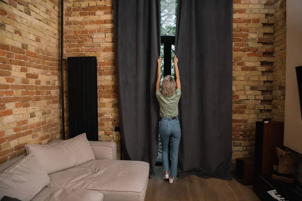 Visão traseira da jovem mulher em jeans azul cortinas de janela de abertura no quarto — Fotografia de Stock