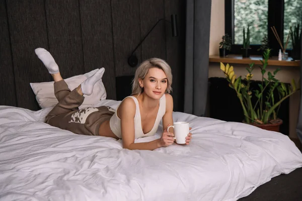 Schöne junge Frau blickt in die Kamera, während sie mit einer Tasse Tee im Bett liegt — Stockfoto